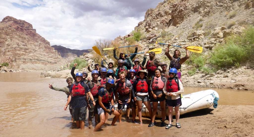 A group of people wearing life jackets stand in shallow water beside a raft. Many of them raise their arms or oars in the air in celebration. 